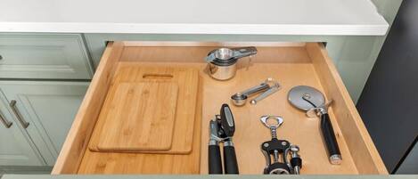 A kitchen drawer with a wooden cutting board, a metal measuring cup, a can opener, a bottle opener, and a pair of scissors.