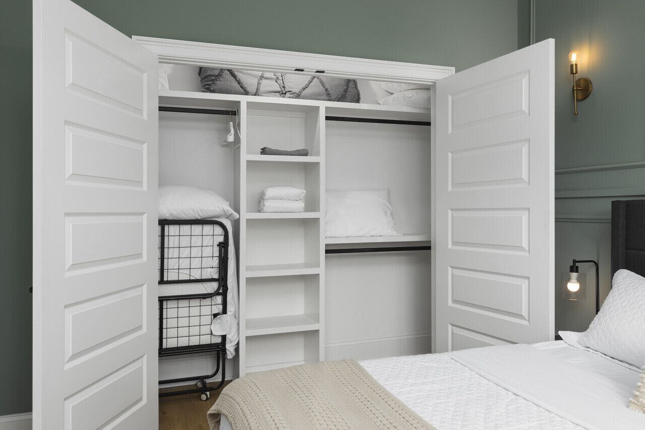 Une chambre à coucher avec un lit, une armoire blanche dont les portes sont ouvertes, et une table de chevet avec une lampe.