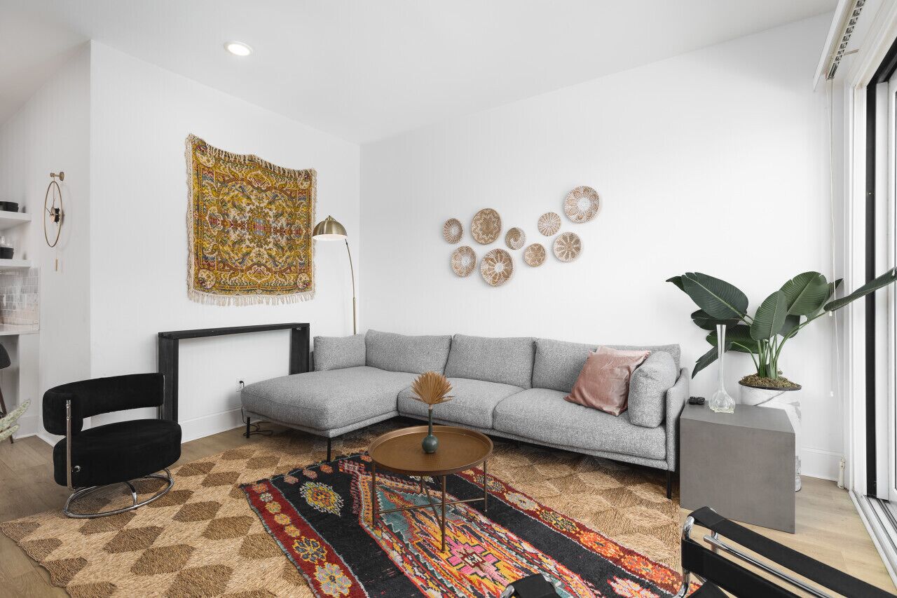 A modern living room with a grey sectional sofa, a black chair, a patterned rug, and a large tapestry on the wall.