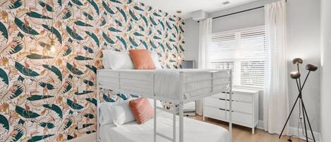 Una cama litera con dos almohadas, un escritorio blanco, una lámpara de pie y una ventana con persianas.
