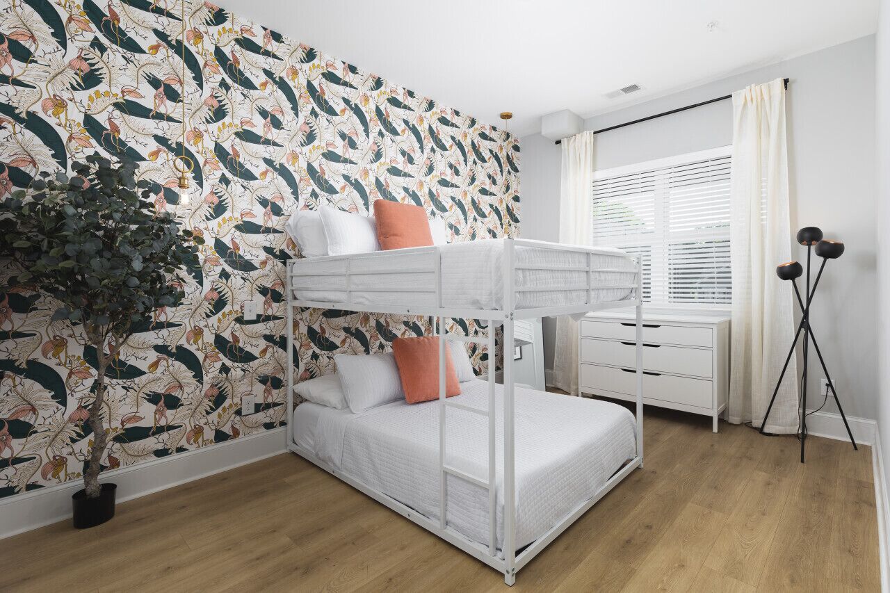 A bunk bed room with a patterned wallpaper, a potted plant, a bedside table, and a lamp.