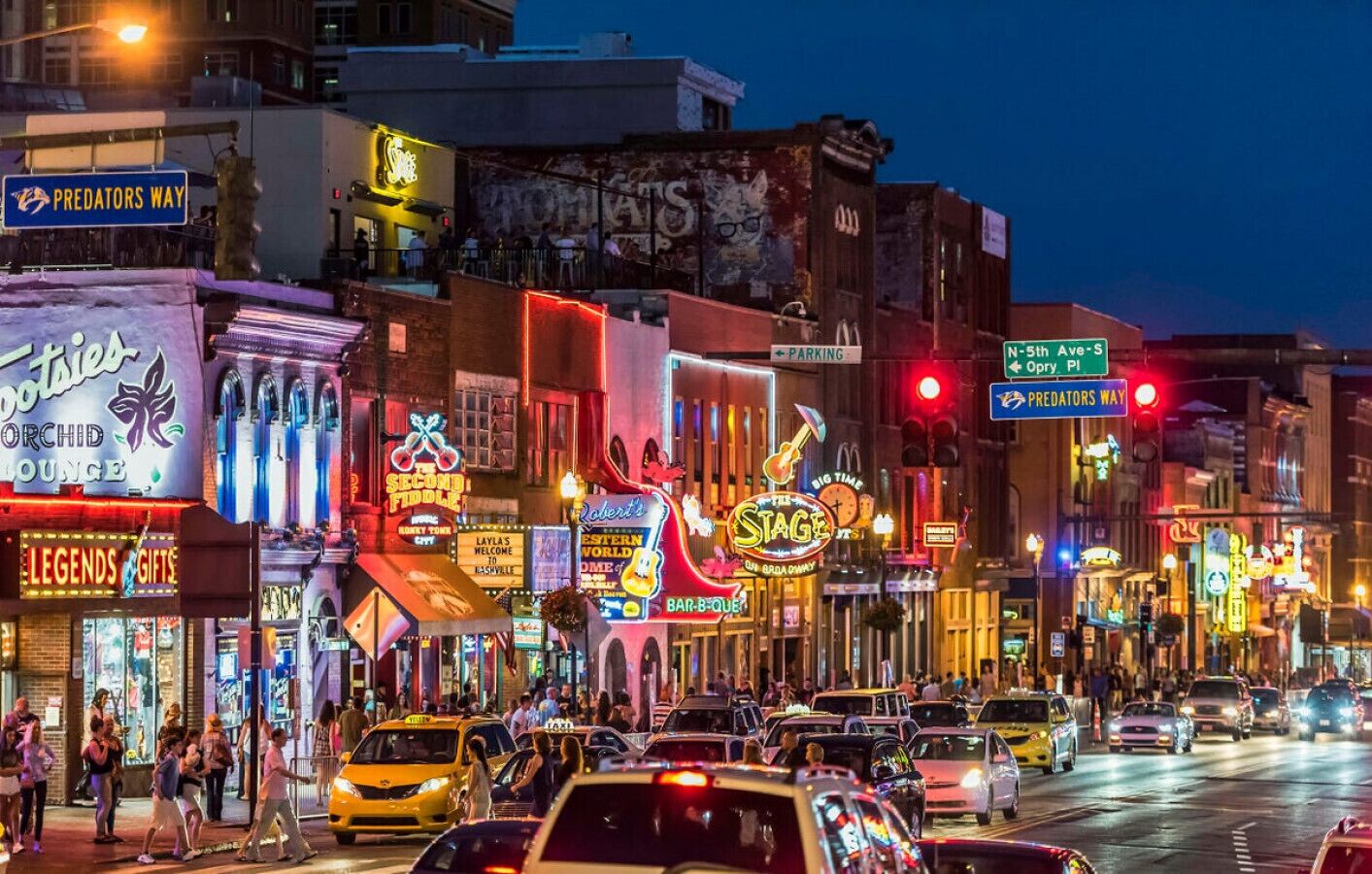 A busy urban street at night with various illuminated storefronts and signs, including a prominent 'Predators Way' sign.