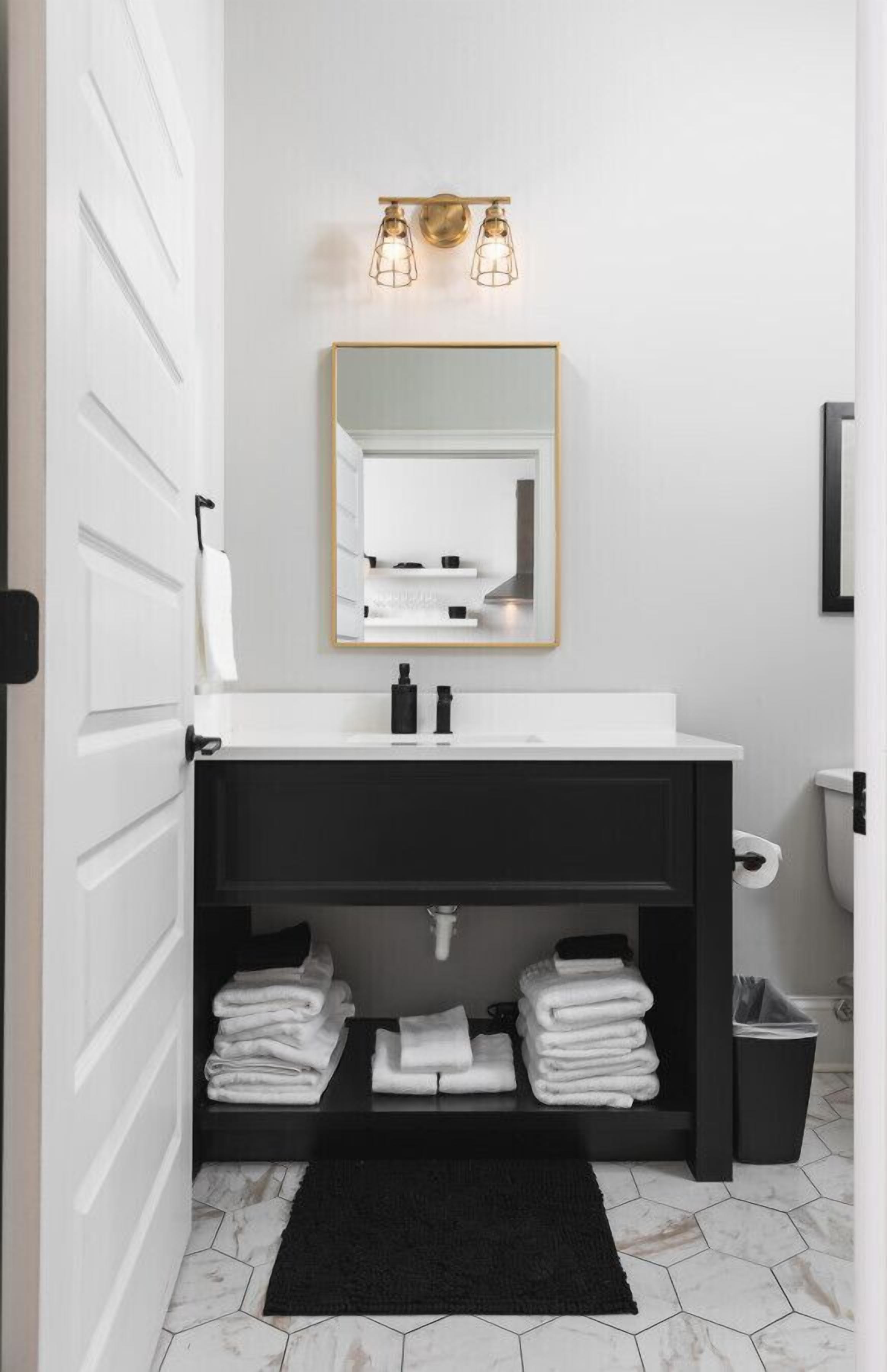 Een moderne badkamer met een witte wastafel, een zwart aanrechtmeubel en een spiegel met gouden lijst. Er liggen handdoeken op het aanrechtmeubel en een zwart vloerkleed op de vloer.