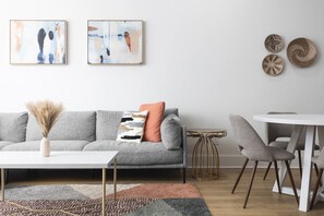 Un salon moderne avec un canapé gris, une table basse blanche et une petite table à manger avec des chaises. Deux tableaux abstraits ornent le mur, et trois paniers en osier sont disposés dans la pièce.