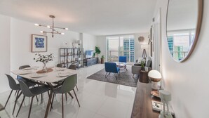 A modern living room with a dining area, a large window, and a view of the cityscape. - The Club- RE Rentals (Miami)
