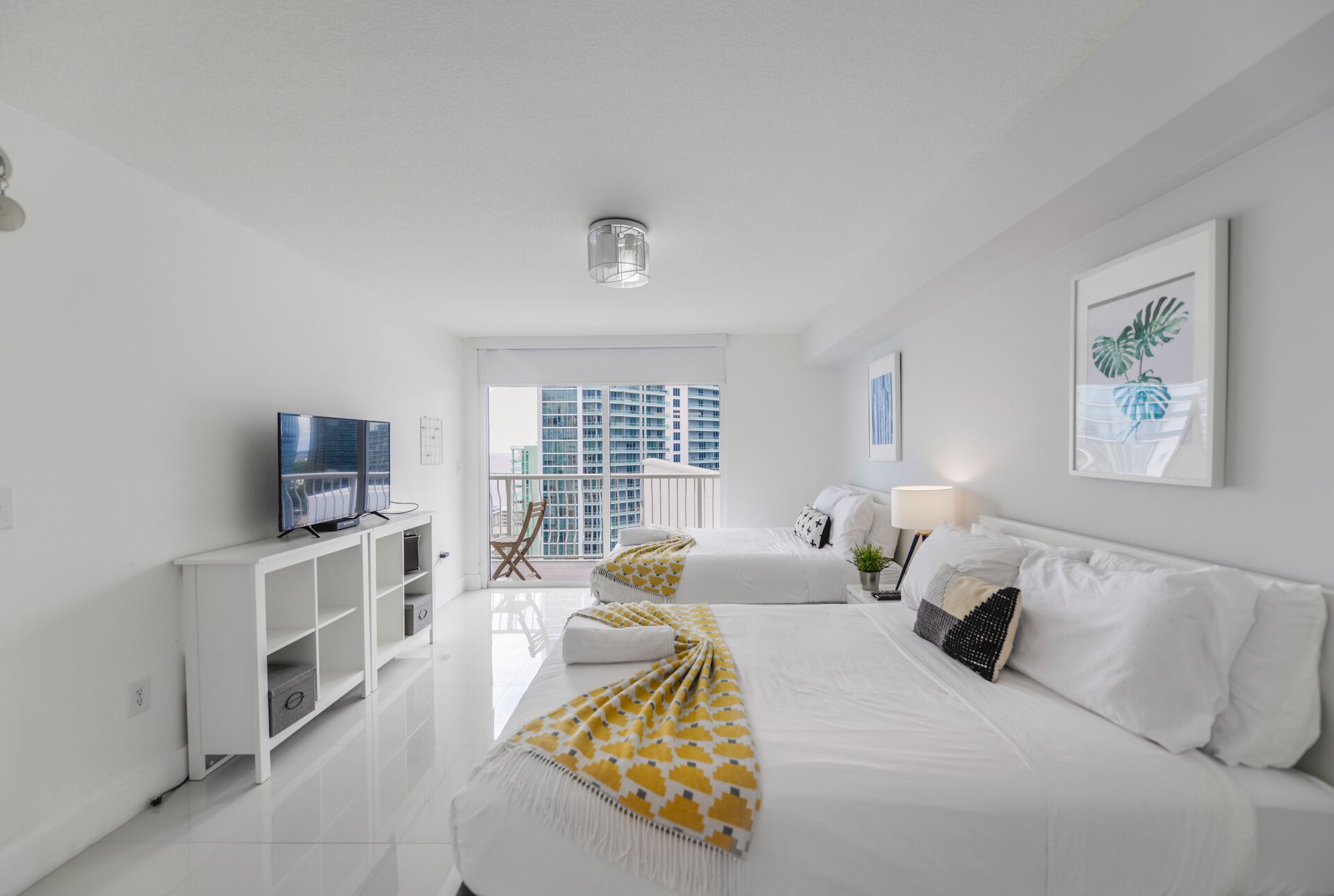 Une chambre d’hôtel moderne dotée d’un grand lit, d’une télévision et d’un balcon offrant une vue sur la ville, le tout dans des tons de blanc.
