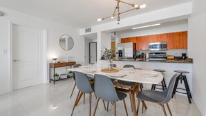 A modern kitchen with stainless steel appliances, wooden cabinets, and a dining area with a white table and grey chairs. - The Club- RE Rentals (Miami)