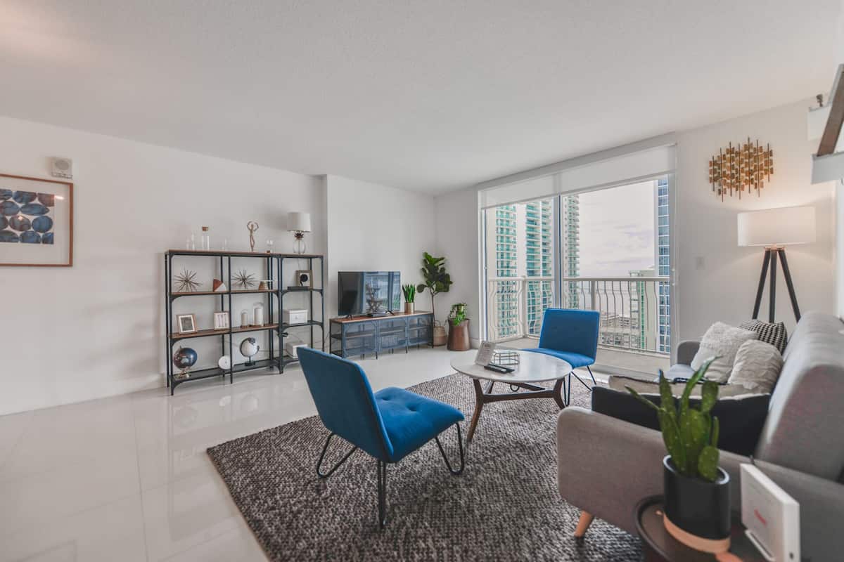 Uma sala de estar moderna com uma poltrona azul, um sofá cinzento e uma mesa de centro. Há uma estante com diversos objetos decorativos e uma janela ampla com vista para edifícios altos.