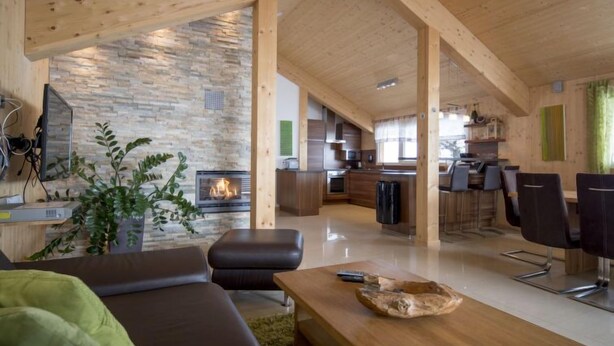 A modern interior with a stone fireplace, wooden beams, a living area with a sofa and coffee table, a kitchen, and a dining area with chairs.