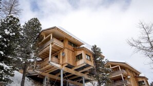 A wooden chalet with multiple balconies, built on stilts, surrounded by snow-covered trees.