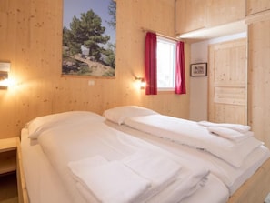 Quarto com duas camas, paredes de madeira, janela com cortinas vermelhas e um quadro de uma árvore.