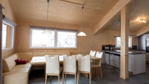 Ein modernes Interieur mit Holzbalkendecke, einem Essbereich mit weißen Stühlen, einer Küchenzeile und großen Fenstern, die für natürliches Licht sorgen.