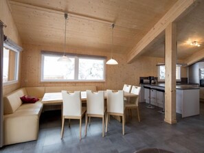 Un interior moderno con techo de madera, un área de comedor con sillas blancas, una zona de cocina y amplias ventanas que ofrecen luz natural.