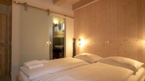 A room with two beds, wooden walls, a bathroom visible through an open door, and a wall-mounted light fixture.