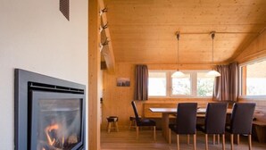 A cozy cabin interior with a fireplace, wooden dining table, chairs, and a window with curtains.