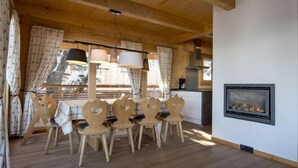 Ein Essbereich mit Holztisch und -stühlen, einem Kamin und einer durchgehenden Tür erkennbaren Küche.