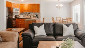 A modern living room with a black leather sofa, wooden dining table, and a kitchen area with wooden cabinets and a stone backsplash.