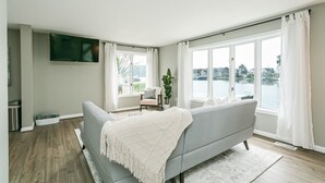 Una sala de estar moderna con un sofá gris, un televisor de pantalla plana montado en la pared y ventanas amplias que ofrecen vistas al agua.
