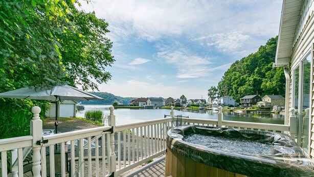 Una terraza con jacuzzi, un banco de madera y una baranda blanca con vista a un lago y casas al fondo.