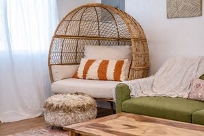Una silla de mimbre con cojín, un sofá verde, una mesa de centro de madera y un otomano mullido en una sala de estar.