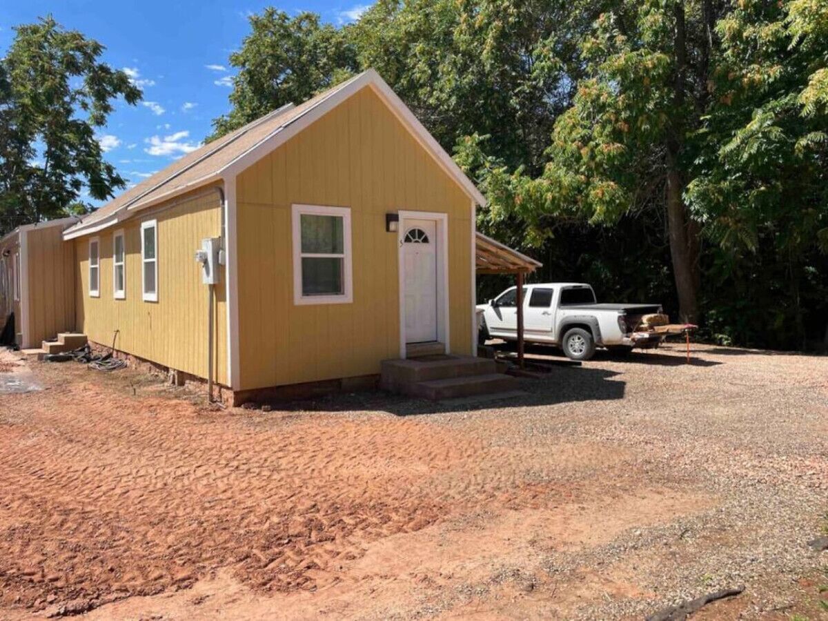 A small yellow building with a white door and windows, a gravel driveway, and a white pickup truck parked nearby.