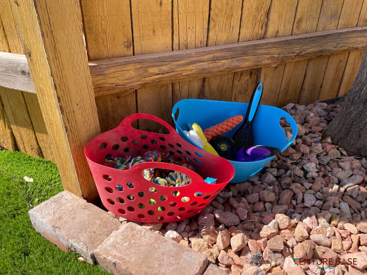 Two plastic baskets filled with various items, placed on a gravel surface near a wooden fence.