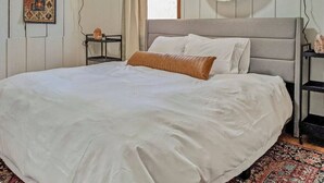 Een groot bed met witte lakens, een grijs hoofdeinde en een bruin sierkussen. Daarboven hangt een wanddecoratie van geweven materiaal, een klein plankje met een zoutlamp, en op de vloer ligt een vloerkleed met een patroon.