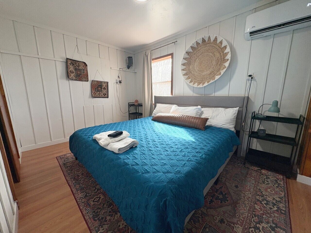 A bedroom with a bed, a wall-mounted air conditioner, a bedside table with a lamp, and a patterned rug.