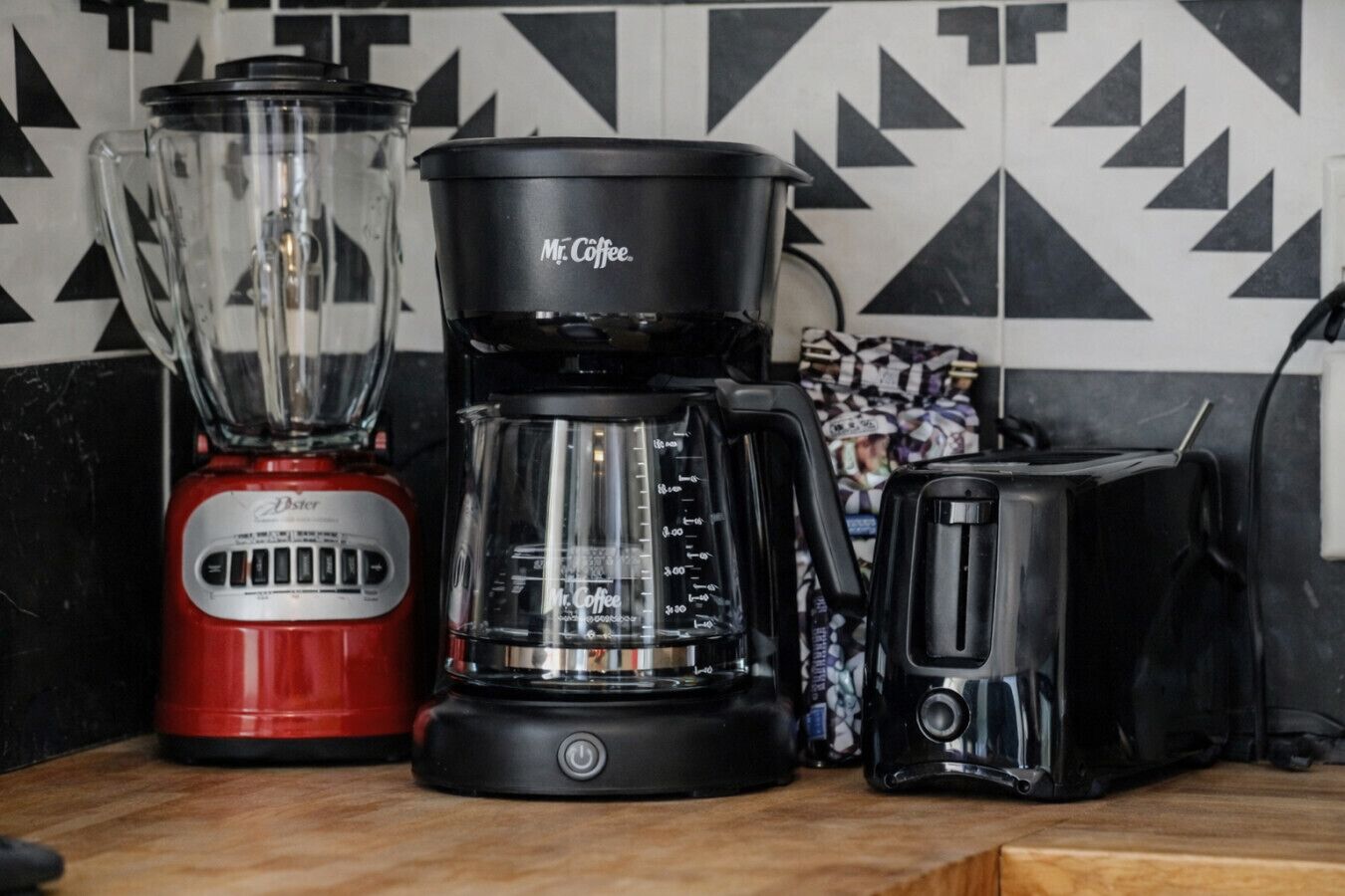 Una encimera de cocina con una licuadora roja Oster, una cafetera Mr. Coffee y un tostador negro.