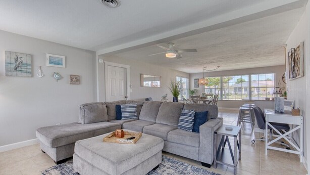 A spacious living room with a large gray sectional sofa, a small ottoman, a dining area with a table and chairs, and a kitchen area with a white countertop and appliances. - Cresting Wave Cottage by the Sea (Ormond Beach)