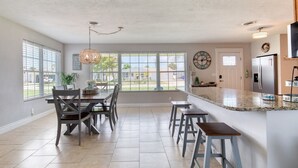 A spacious kitchen with a large island, a dining area with a table and chairs, and a view through large windows. - Cresting Wave Cottage by the Sea (Ormond Beach)