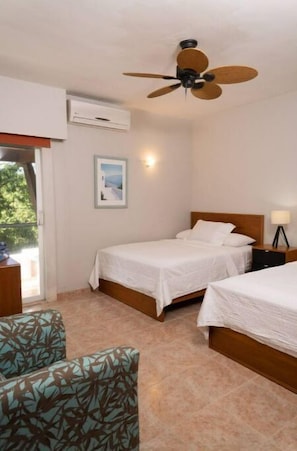 A hotel room with two single beds, a ceiling fan, an air conditioning unit, a framed picture on the wall, and a view of greenery outside.