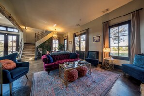 A spacious living room with a staircase, large windows, and a chandelier.
