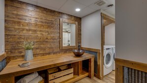 Une coiffeuse en bois avec un lavabo, un miroir et un vase rempli de fleurs. On aperçoit une machine à laver au fond.