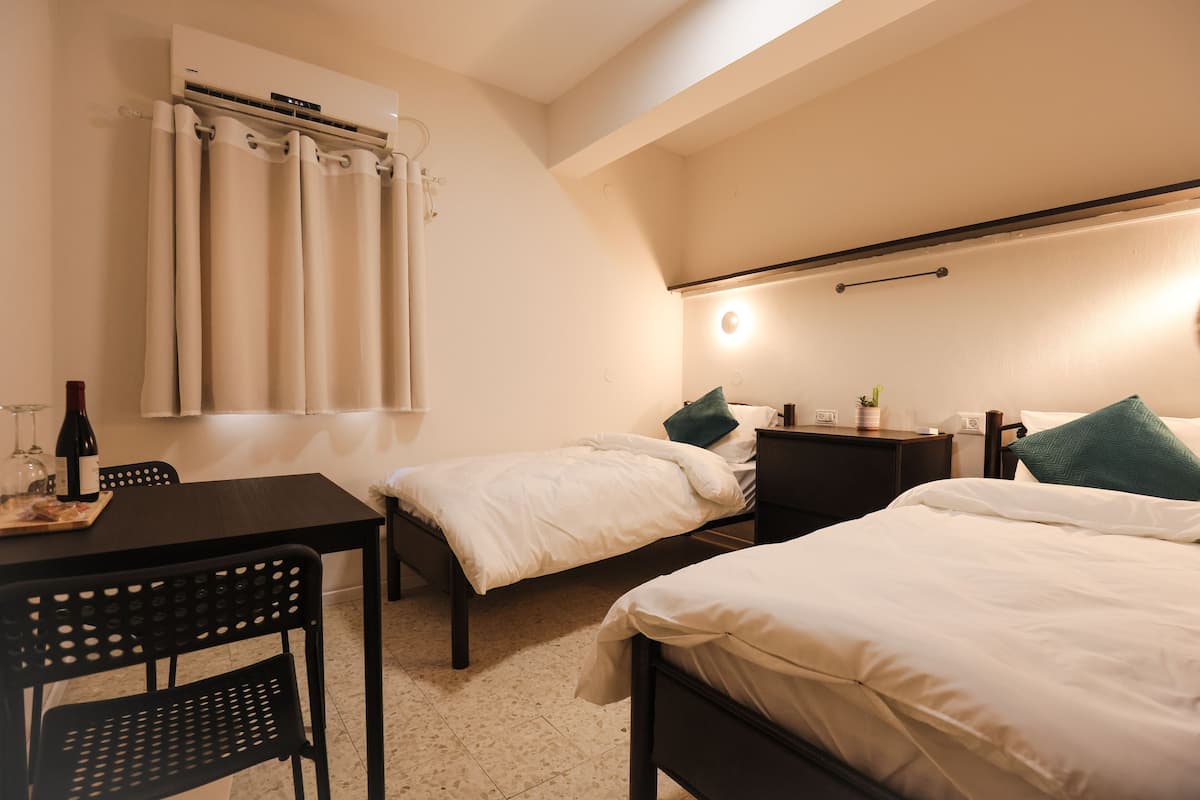 A hotel room with two beds, a dining table, chairs, a small table with a bottle, and a wall-mounted air conditioner.