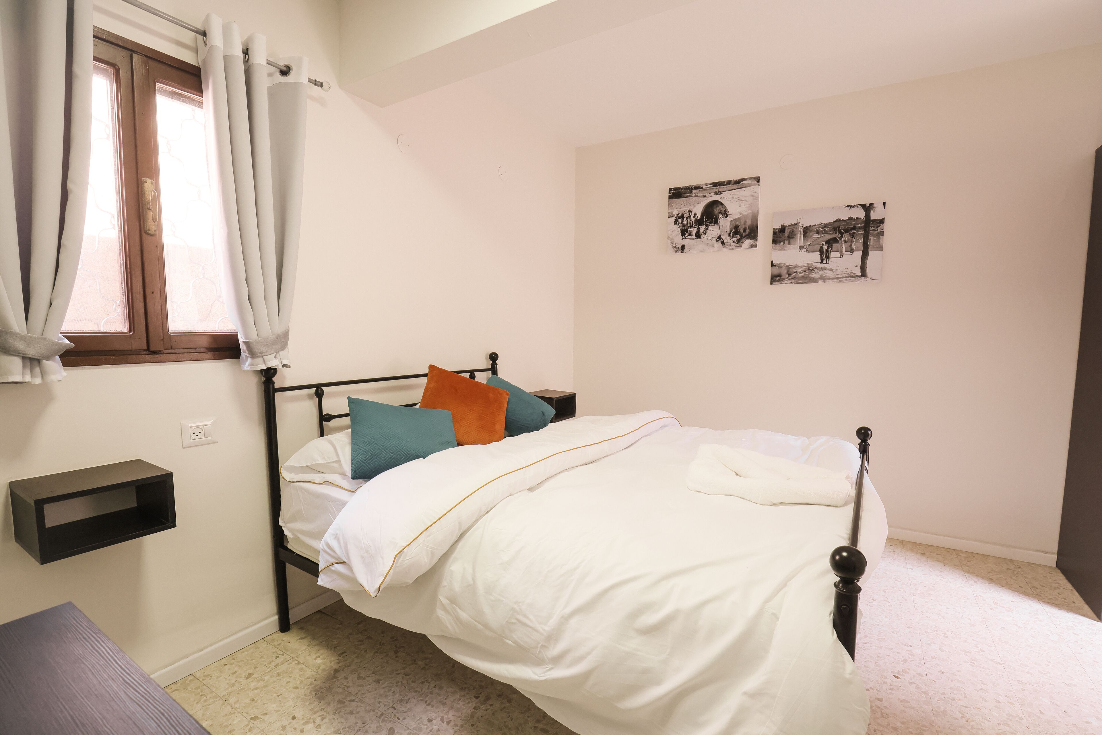 A bedroom with a bed, a bedside table, a shelf, and a window with curtains.