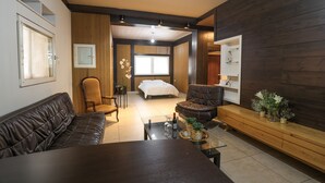 Een moderne woonkamer met een bank, fauteuil en salontafel, een slaapkamer met een bed en raam, en een houten kastje met decoratieve objecten.