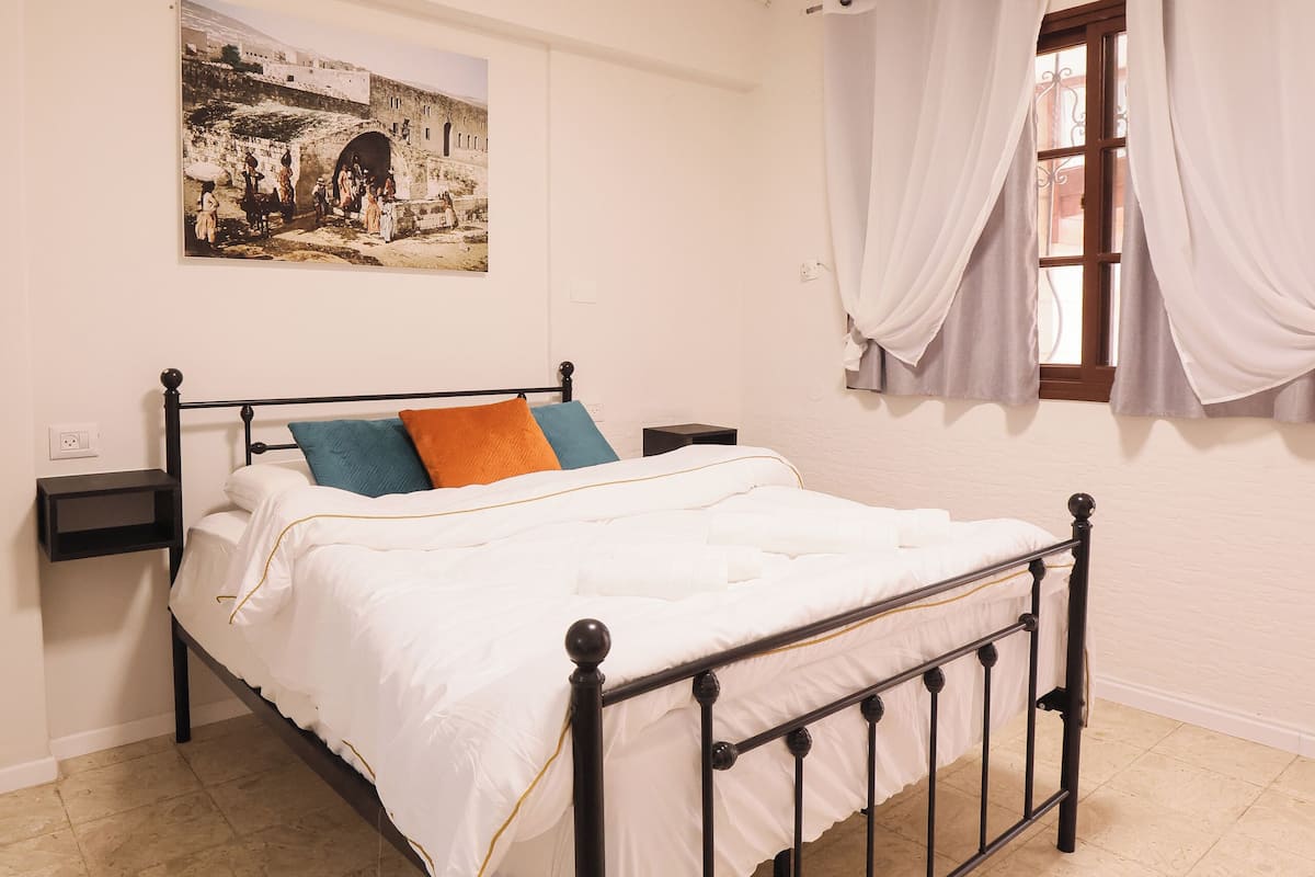 A neatly arranged bedroom with a bed, a nightstand, a window with curtains, and a wall-mounted picture.