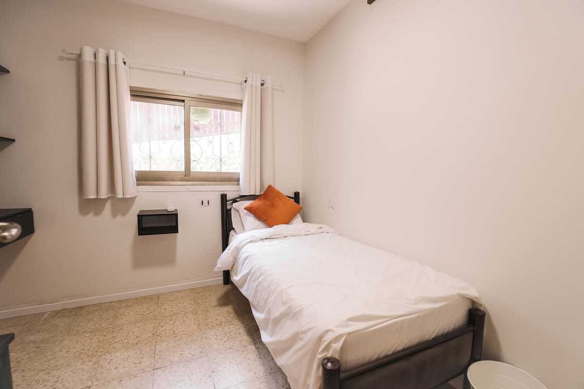 A small, clean room with a single bed, a window with curtains, a small shelf, and a radiator.