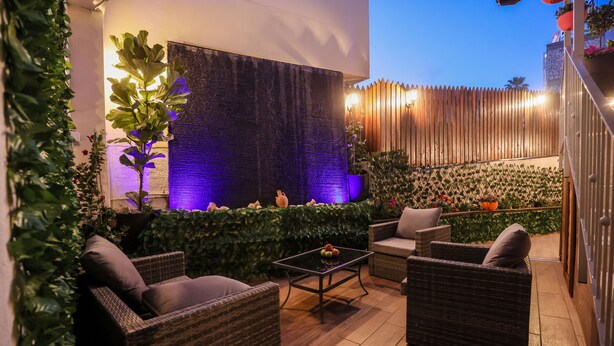 Een terras buiten met wicker meubels, een tafel met fruit en een decoratieve muur met een waterpartij.