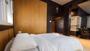 Een tweepersoonsbed met witte lakens, een houten paneel, een kruisbeeld en een bruine lederen fauteuil in een kamer met betegelde vloer.