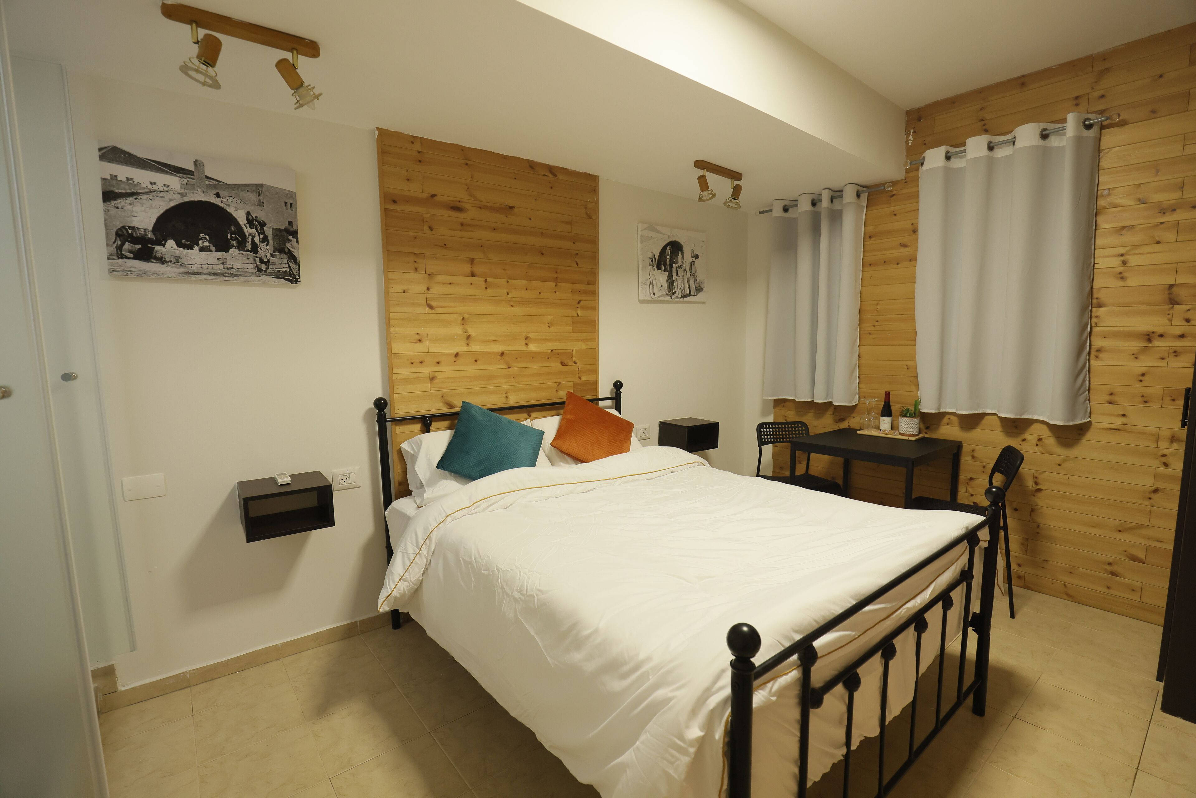 A bedroom with a wooden headboard, a bed with white bedding, two nightstands, a small table with a chair, and a window with curtains.