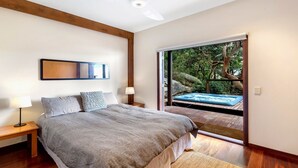 A bedroom with a large bed, bedside lamps, a wooden floor, a ceiling fan, and a sliding glass door leading to an outdoor hot tub area. - Atlas - House for all seasons, spa with ocean views and cozy fireplace (KILLCARE)