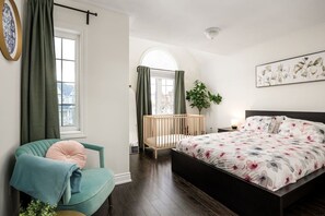 Ein Schlafzimmer mit einem Bett, einem Babybett, einem Sessel, einem Fenster und einer Pflanze.