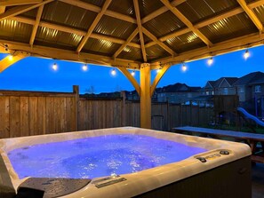 Ein Whirlpool unter einer Holz-Pergola mit Lichterketten, umgeben von einem Holzzaun und Wohnhäusern im Hintergrund.