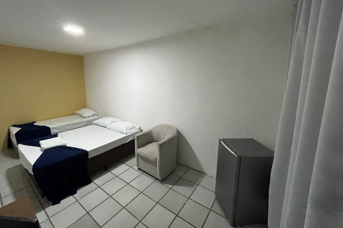 A small, single-room hotel room with a bed, a chair, a desk, and a refrigerator.