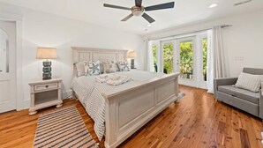 Ein Schlafzimmer mit einem großen Bett, einem Deckenventilator, einem Nachttisch, einer Lampe, einem grauen Sofa und Blick auf Grün durch das Fenster.