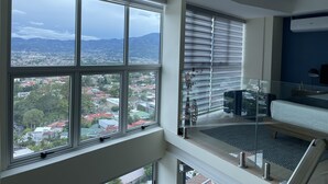 Ein modernes Hotelzimmer mit großen Fenstern und Stadtblick, einem Balkon mit Glasgeländer sowie einer Sitzecke mit Sofa und kleinem Tisch.