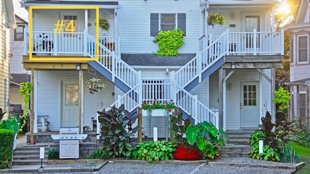Una casa de dos pisos con un porche delantero, una escalera y un pequeño jardín con plantas y una parrilla.