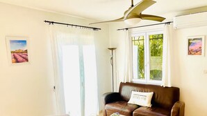 A living room with a brown sofa, two white armchairs, a glass coffee table, a ceiling fan, and a window with curtains.